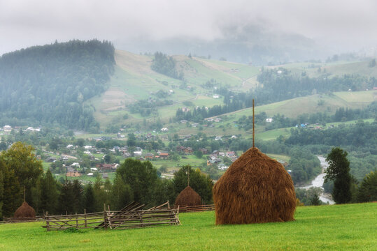 Idylic foggy rural mountain landscape in Carpathians. Traditional haystacks standing on a green pasture overlooking misty  hills  covered with forest and rural settlements in the distant background.