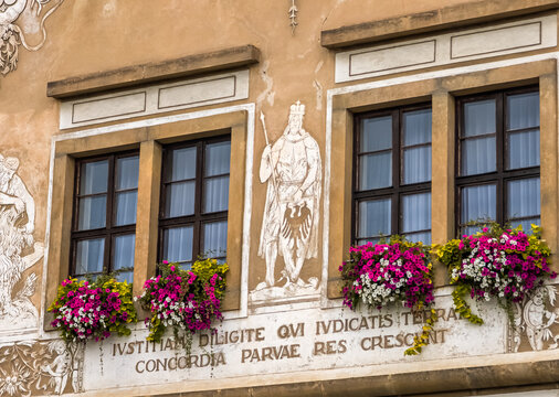 Detailed view of ancient sgraffito wall art on a historical building featuring a figure with a shield and Latin inscriptions below windows with pink flowers.