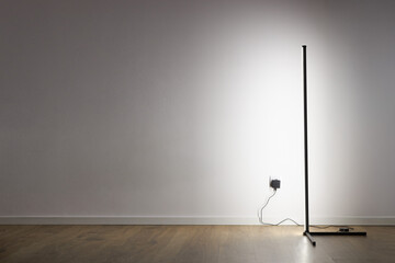 Minimalist LED floor lamp illuminating white wall in modern interior with wooden floor and visible power outlet