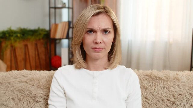 Middle-aged angry woman sits on home sofa shaking head firmly, expressing clear refusal or disapproval. Adult sad lady reacts with seriousness, showing personal boundary and emotional confidence.