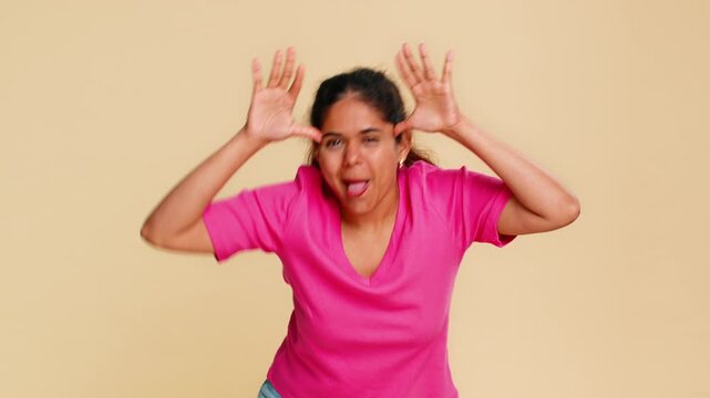 Indian woman playfully sticks tongue out towards camera, cheerfully expressing humor fun. Girl on beige background engaging in playful teasing, showing carefree, lively attitude with funny gesture