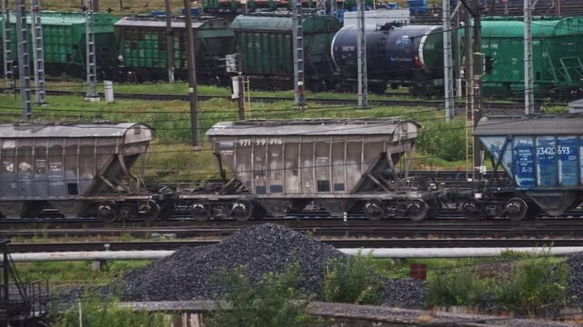 A wide shot of a busy rail yard with numerous freight trains and hopper cars on the tracks. Piles of bulk material, possibly coal or gravel, sit in the foreground of the industrial setting