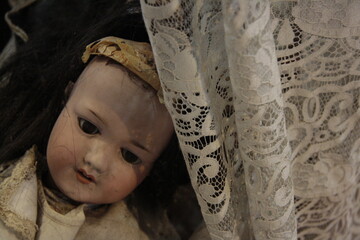 Vintage photo of an old ceramic doll behind a white lace curtain. Dim lighting creates a slightly eerie and nostalgic atmosphere, evoking childhood memories and timeless melancholy.
