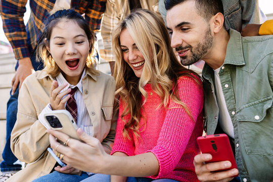 Group of diverse young friends outdoors using smartphones, laughing together while watching entertaining social media content. Friendship, youth community and technology lifestyle concept. - Powered by Adobe