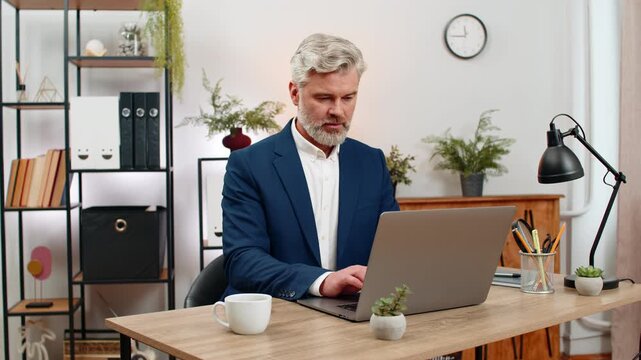 Middle-aged businessman at home office opens laptop ready to boost output starting productive workflow. Freelancer guy at table types meeting work deadlines tracking progress boosting output daily up.