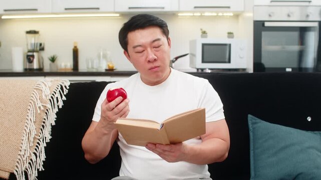 Asian man at home bites red apple enjoys juicy taste chews slow and smiles feeling fresh and awake. Chinese guy on sofa reads health tips in book marks page then eats more and feels fine and calm good