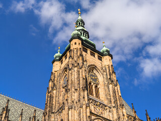 Fototapeta premium Close up of a magnificent cathedral clock tower with green spires and golden details reaching into a vibrant blue sky with soft white clouds.