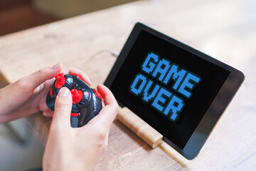 person grips a game controller in hands while a tablet screen displays the words GAME OVER