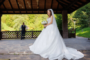 Fototapeta premium Bride in a white gown stands proudly while groom looks on in a beautiful outdoor setting