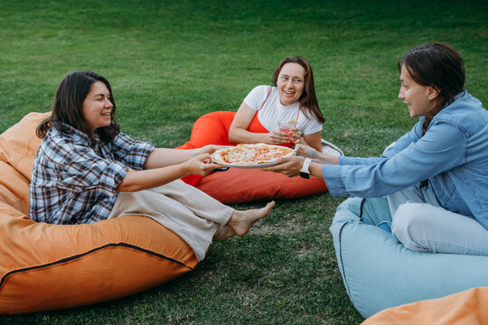Friends relaxing on colorful bean bags outdoors sharing food and drinks in casual social gathering lifestyle moment with small dog. Woman informal communication during dinner with pizza in backyard 