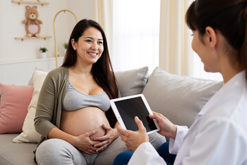 Smiling pregnant woman having consultation with female doctor using digital tablet in clinic. Obstetrician providing prenatal care, representing modern healthcare, pregnancy, technology in medicine
