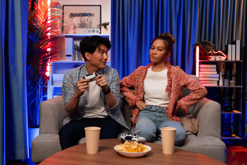 Sitting sofa of happy couple playing video game with joysticks, smiling man playing challenge, raising fist up to be winner the challenge at modern neon blub light living room at home. Infobahn.