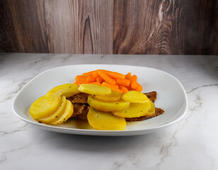 A plate features tender slices of beef alongside vibrant yellow potatoes and bright orange carrots. The meal is presented on a smooth white plate, set against a wooden backdrop