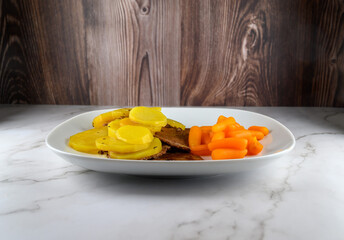 A plate features tender slices of beef alongside vibrant yellow potatoes and bright orange carrots. The meal is presented on a smooth white plate, set against a wooden backdrop