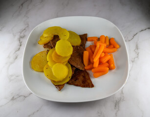 A plate features tender slices of beef alongside vibrant yellow potatoes and bright orange carrots. The meal is presented on a smooth white plate, set against a wooden backdrop