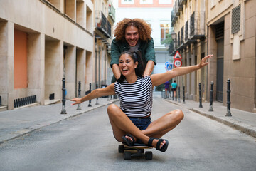 Young woman sitting on skateboard with open arms while man pushing her in urban street