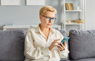Mature confident woman pensioner sitting on sofa in living room at home using smartphone for social...