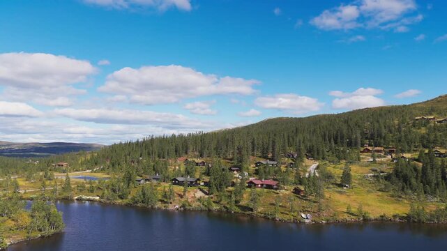Slow Aerial Pan Shot Revealing a Picturesque Norwegian Mountain Cabin Village, Dense Pine Forest, and Small Lake Under a Blue Sky