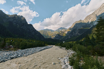 Pristine mountain landscape with a sandy riverbank, scattered stones, and distant rugged peaks beneath a bright sky. The scene conveys calm, adventure, and natural beauty.