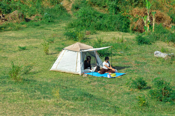 Sitting side by side on a picnic mat near their tent, two teenage girls rest calmly, looking toward something unseen, sharing a serene pause in their outdoor day.
