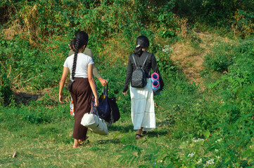 With backpacks and a tent bag in hand, three girls walk side by side along a grassy, sunlit path leading away from the campsite, signaling the peaceful end of their outdoor trip.