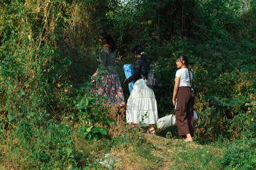 The girls stop mid-path, gazing at the distant landscape with smiles, appreciating the natural beauty while carrying their bags and tent under bright sunlight.