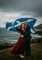 A couple with red hair holds the flag of Scotland on a windy coastal cliff. Romantic portrait celebrating Scottish heritage