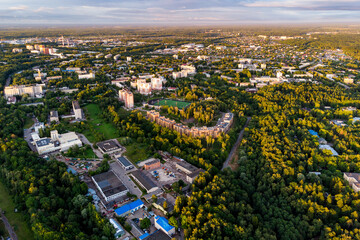 Aerial view of a green city with modern residential buildings, a stadium, and vast forests under a golden hour sky. Urban landscape surrounded by nature