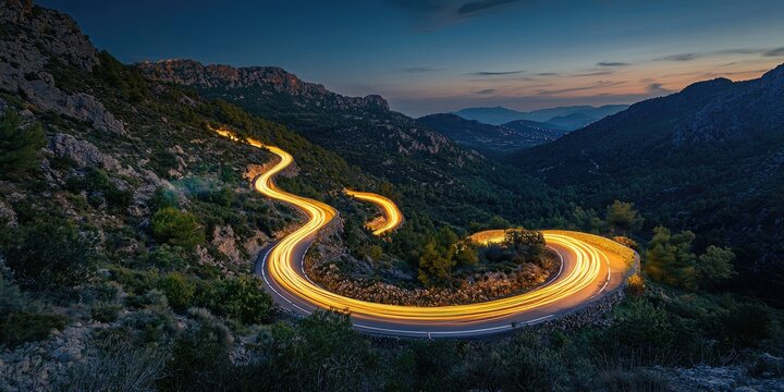 A winding mountain road at dusk with a curving light trail, surrounded by lush greenery and towering mountains in the background.