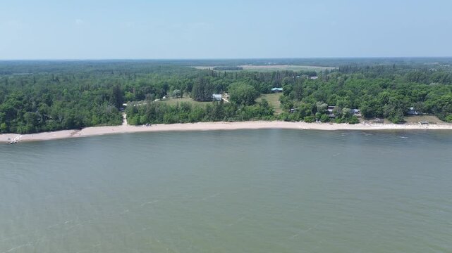This cinematic drone footage captures an expansive view of a large waterway and its undeveloped shoreline. The calm water reflects the sky, bordering the endless green forest on a summer day.