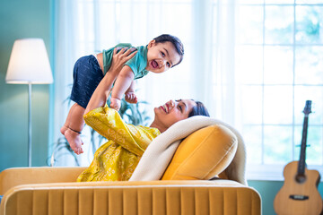 Happy Indian mother catching her baby boy on sofa, enjoying playful bonding time
