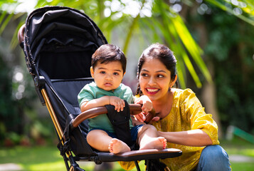 Indian Mom walking infant in stroller through sunny garden, sharing loving and peaceful moments