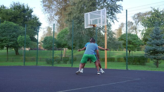 Active fit African American male streetball player performing step back shot, scoring field goals while black athletes playing half court basketball game in morning outdoors.