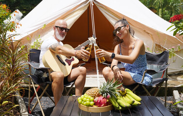 A man and woman is joyfully toasting with beer near a cozy tent at a glamping site.