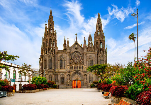 Front view of the Church of San Juan Bautista in Arucas, Gran Canaria, with ornate portal, palm trees, bushes, benches and large open square.