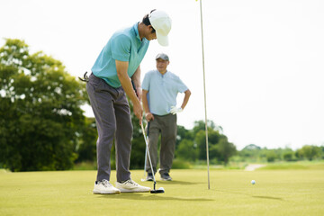 Asian golfer putting on green with senior cheering nearby.