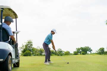 Young Asian golfer hitting shot while senior watches from golf cart.