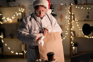 Young female doctor in eyeglasses and Santa Claus hat on the shift unpacking takeaway food in paper bag working at night on Christmas Eve in hospital. Holidays, medicine and consumption