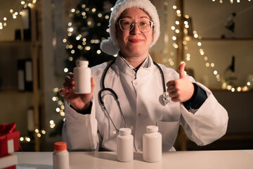 Concept of medicine, healthcare and holidays. Young female doctor in Santa hat recommended pills bottle pointing thumb-up in ok good gesture while working on night shift on Christmas Eve