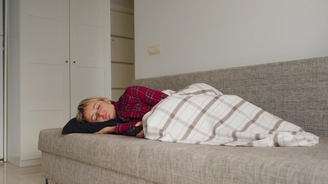 Young woman lies curled on a sofa in red plaid pajamas, holding her lower abdomen in visible discomfort. The TV remote is nearby. Concept of menstrual pain and rest during periods. Natural light, mid
