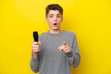 Singer Teenager man picking up a microphone isolated on yellow background surprised and pointing front