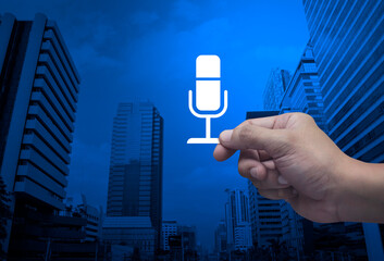 Hand holding microphone flat icon in fingers over modern office city tower and skyscraper, Technology communication concept