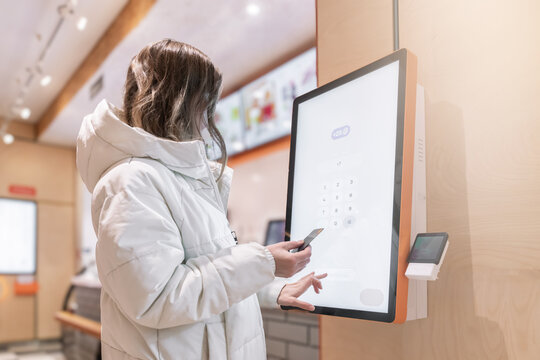 A woman in a white jacket pays for the purchase of an order in a fast food restaurant via a touchscreen with her own credit card. Enter the phone number on the screen so that the receipt is sent to