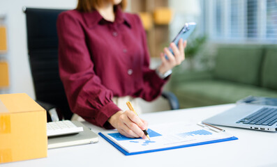 Asian entrepreneur woman using clipboard in home office. Surrounded by boxes, laptop & tablet. ecommerce, SME, delivery and startup business themes.