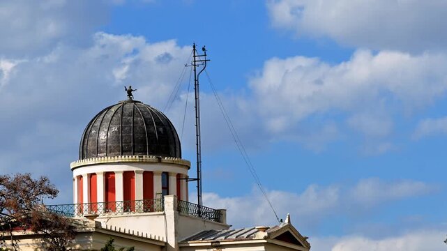 Time lapse footage shows clouds passing by at blue sky behind a building's dome with a lightning rod and wind vane attached at the small porch.