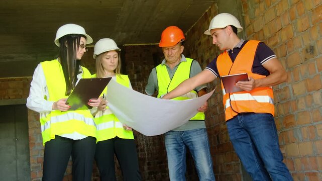 Builder points to fragment of floor plan standing near colleagues. Construction worker tells engineers and partner about specifics of layout