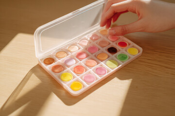 Naklejka premium Hand arranging watercolor palette on wooden table, sunlight casting soft shadows, vibrant paint colors, creative art supplies, warm atmosphere, artistic inspiration, close up view