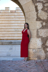 A beautiful teenage girl with long dark hair, dressed in red and wearing high heels, leaning under a Roman archway on a street in a typical Andalusian town. In the background is a wall.