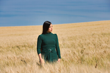 A beautiful, dark-haired teenage Latin girl dressed in green stands among a dry, yellow wheat field on a summer day. In the background, the blue sky.