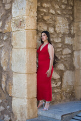 A beautiful teenage girl with long dark hair, dressed in red and wearing high heels, leaning under a Roman archway on a street in a typical Andalusian town. In the background is a wall.